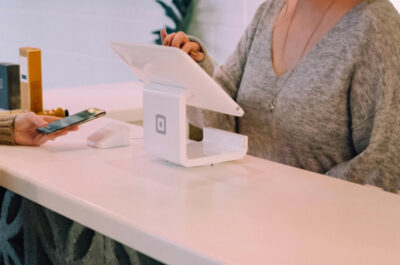 Zwei Frauen stehen sich an einem modernen weißen Tresen gegenüber. Die Frau auf der einen Seite des Tresens hält ein Smartphone in der Hand und ist im Gespräch mit der anderen Frau, die neben einer weißen Point-of-Sale-Station steht. Die Umgebung wirkt einladend und modern.