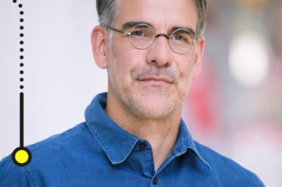 Das Bild zeigt einen Mann mittleren Alters mit graumelierten Haaren und einem gepflegten Bart. Er trägt eine runde Brille und ein blaues Hemd. Der Hintergrund ist verschwommen, wodurch der Fokus auf dem Mann liegt. In der oberen linken Ecke befindet sich ein Logo mit der Aufschrift "brand eins Netzwerk".
