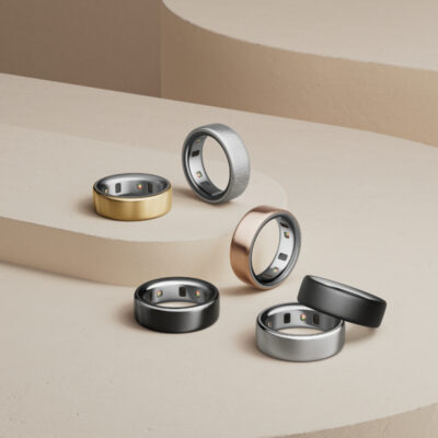 Ein stilvoll arrangiertes Set aus sechs modernen Smart-Ringen in verschiedenen Metallfarben – darunter Gold, Schwarz, Silber, Roségold und gebürstetes Grau – auf einer minimalistischen beigen Stufenplattform. Die Ringe sind mit eingebauten Sensoren auf der Innenseite ausgestattet und reflektieren einen eleganten, technologischen Look. Die Komposition betont die luxuriöse Verarbeitung und die Vielseitigkeit der Smart-Ringe als modische Wearables.